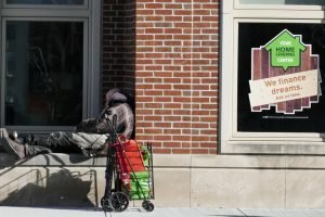 Unhoused man sits in front of a bank offering home loans.